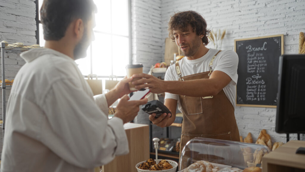 Man paying barista with mobile phone in bakery while holding coffee indoors representing hospitality