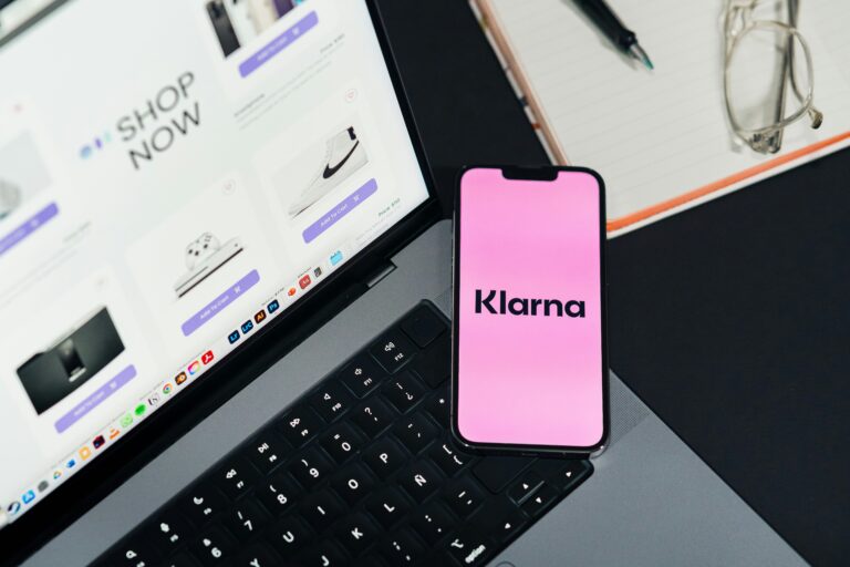 Klarna on phone screen. Top view of workspace. Table desk with laptop with ecommerce on screen